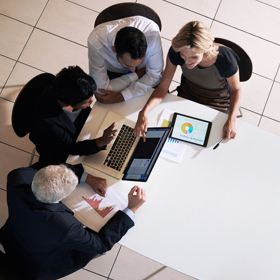 Group of people gathered around table discussing over laptops and tablets